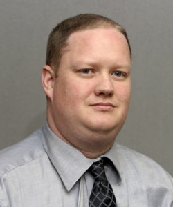 Burton Speakman headshot: SOlomn man with darkblonde hair wearing a light grey shirt and dark tie.