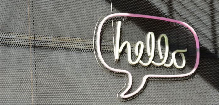 Neon sign in pink and white saying "Hello" inside a chat bubble hangs on a grey metal mesh wall.