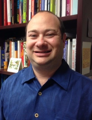 Josh Azriel headshot: Smiling bald man wearing dark blue shirt is in front of a dark wood bookcase.