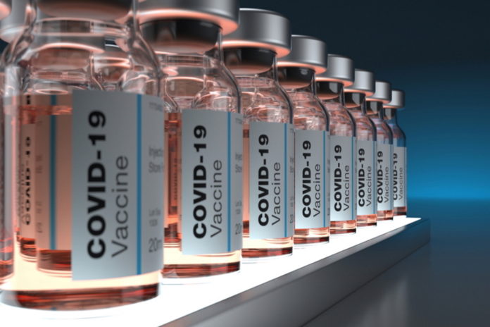 PODCAST COVID-19 vaccines: Clear bottles of the pale pink liquid vaccine labeled and ined up on a shelf in a semi-dark room
