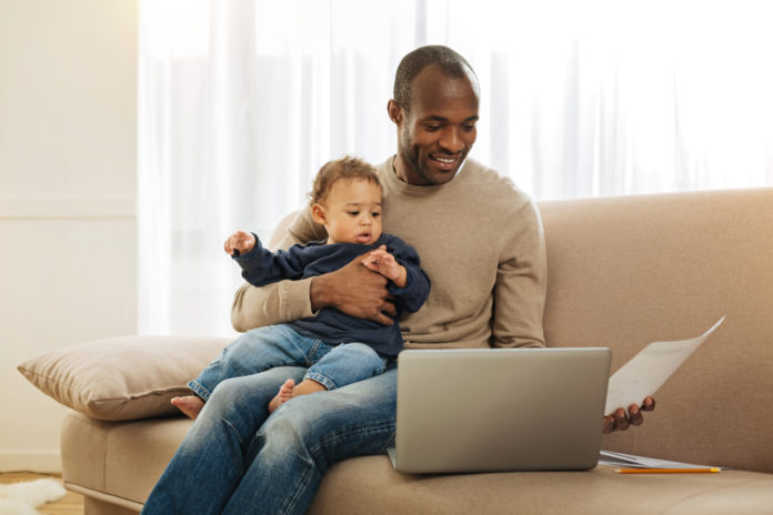 Black man wearing beige sweater sitting on beige couch in front of white curtains holding baby in jeans and navy sweatshirt with laptop on couch