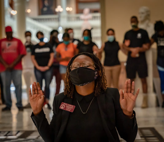 Georgia’s new GOP election law draws criticism, lawsuits Black woman in front of crowd wearing mask, gold wire-rim glasses,and black top with black jacket, hands held up to shoulders eyes closed in reflection