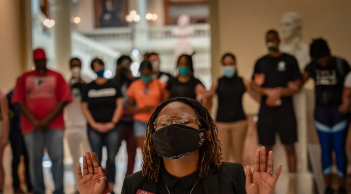 Georgia’s new GOP election law draws criticism, lawsuits Black woman in front of crowd wearing mask, gold wire-rim glasses,and black top with black jacket, hands held up to shoulders eyes closed in reflection
