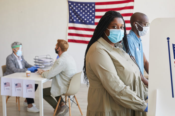 Black woman wearing blue face mask and tan blouse voting with other voters American flag on back wall