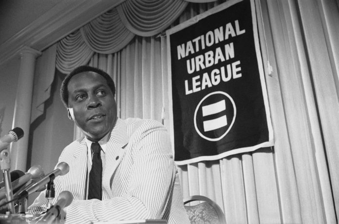 Vernon Jones at press conference in 1977: a Black man wearing white seersucker suit and dark tie standing at podium speaking into microphones