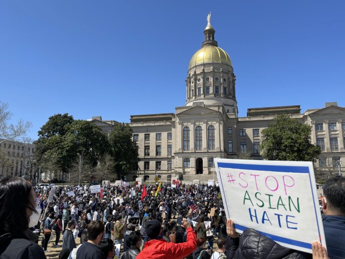 Large crowd of protestors holding signs stand in front of Georgia State Capitol Dome building with white sign in foreground saying #Stop Asian Hate with blue border