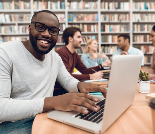 Colleges hold tuition flat for second straight year in Georgia man smiling while on his laptop sitting at light wood table with friends with library book shelves in the background