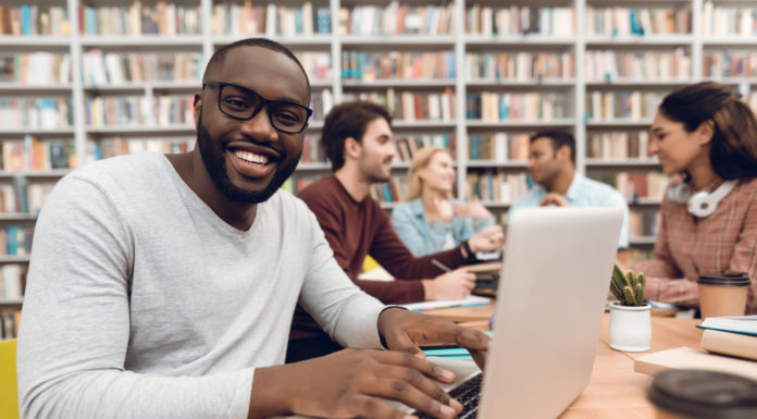 Colleges hold tuition flat for second straight year in Georgia man smiling while on his laptop sitting at light wood table with friends with library book shelves in the background