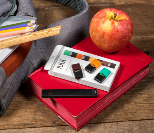 Vapor product curriculum in schools now required by law Gray bookbag on wood desk with vape and a apple beside it on top of a red book