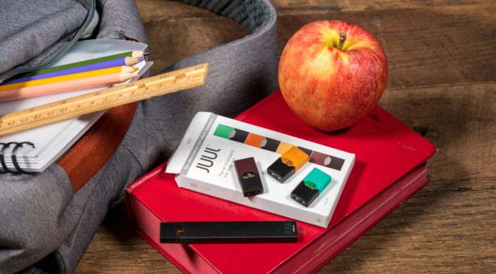 Vapor product curriculum in schools now required by law Gray bookbag on wood desk with vape and a apple beside it on top of a red book