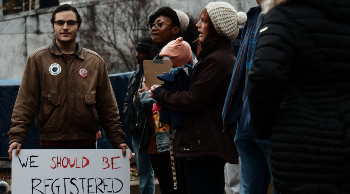 Under pressure, some Georgia corporate leaders slam voting bill Georgia voters standing at outdoor protest- one holding sign saying "We shuld be registered automaticaly"
