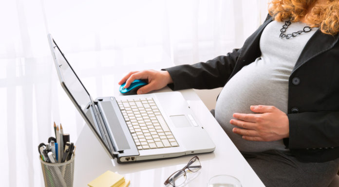 Thousands of state employees to receive paid parental leave Pregnant woman wearing grey top with black jacket holding her belly while sitting at white desk with laptop