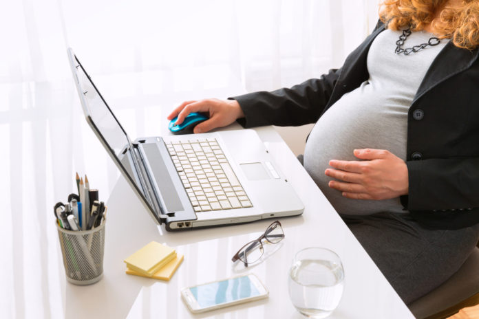 Pregnant woman wearing grey top with black jacket holding her belly while sitting at white desk with laptop