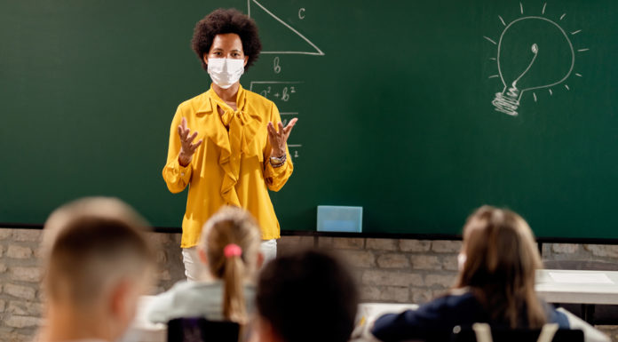 Lessons in Resilience: School staff adapt to COVID-19 changes Black teacher in front of chalkboard with mask on teaching young students