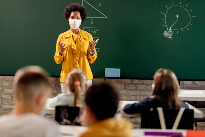 Black teacher in front of chalkboard with mask on teaching young students
