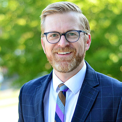 Medicaid Expansion: Craig Wilson headshot - Man with linde hair and beard wearing dark-framed glasses in blue suit with white shirt and blue stripes ties smiling in front of greenery background