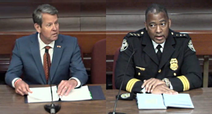 Gov. Brian Kemp white man in gray suit and tie with blonde hair(left) and Chief of Police Todd Coyt (right - black man in navy blue police uniform (right) sit at table with paperwork both facing camera