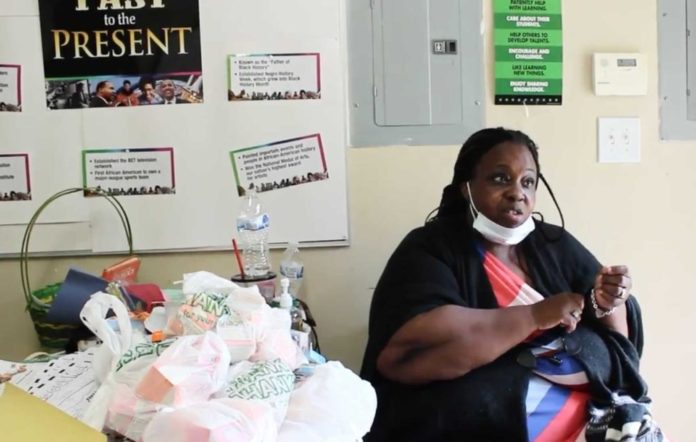 Black woman sitting at table next to food donations with signs on wall behind her