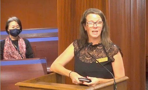 White woman speaks at podium in front of masked woman