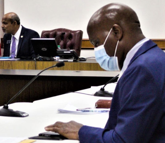 Seeing is believing; students learn faster outdoors Outdoor Learning Study Committee: Bald black man in face mask wearing blue suit sits at table in front of microphone with another black man in suit in the background.