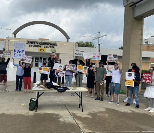 Kennesaw students and faculty rally the call to Mask Up! mask mandate protest for COVID-19 on college campus; masked protestors in group of ~20 hold signs in front of table on rainy day