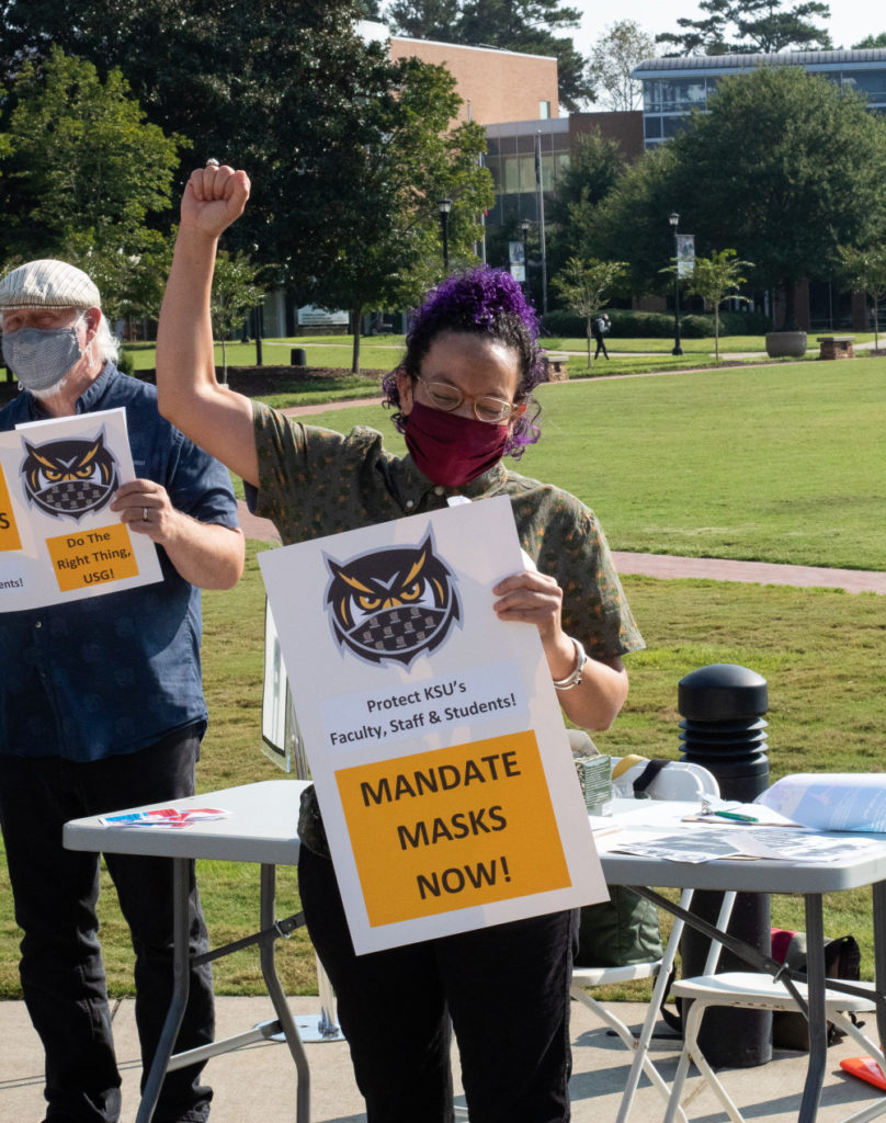 mask mandate protest for COVID-19 on college campus; masked woman with purple hair holds sign declaring 'mandate masks now!'