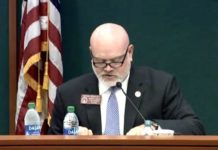 Georgia Medical Cannabis Oversight Committee undecided on testing guidelines Marijuana Oversite Committee: a bald head white man in a dark suit with glasses at a podium with mic and two bottle of water