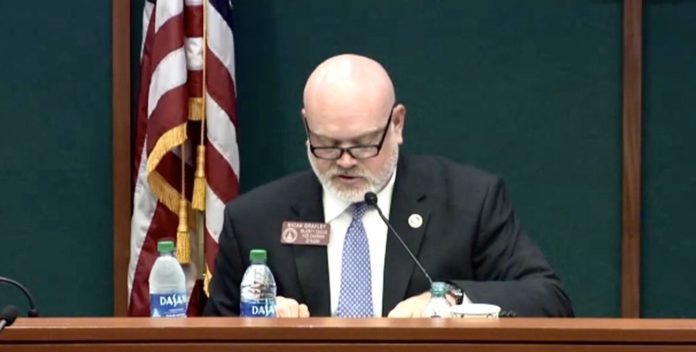 Marijuana Oversite Committee: a bald head white man in a dark suit with glasses at a podium with mic and two bottle of water