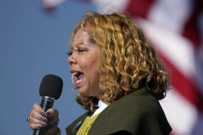 Georgia redistricting: Black woman with curly blonde hair wearing brown jacket and gold top speaking into microphone she is holding in right hand