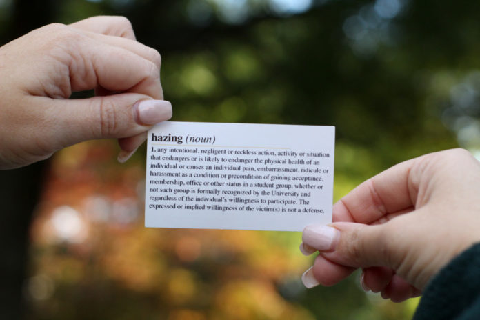Fraternity hazing: Close up of two hands hold white business card with black text that faces camera showing a dictionary definition of hazing