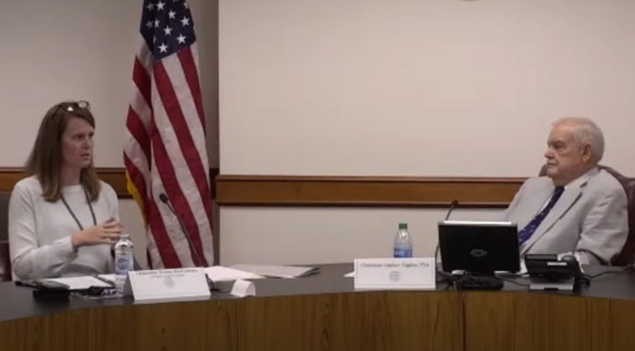 High university fees concern lawmakers Georgia university fees: Man with grey hair in gray suit looking at lady in gray blazer with brown hair at a large brown desk with American flag in the background