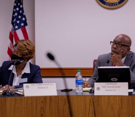 Senate study committee makes recommendations to eliminate food deserts in Georgia Georgia Food Deserts meeting: Bald black man wearing black frame glasses and blue pin-striped suit with left hand on chin to right of a black woman with blond hair in navy suit and white blouse both sit behind brown wood desk in front of American flag and Senate gold seal