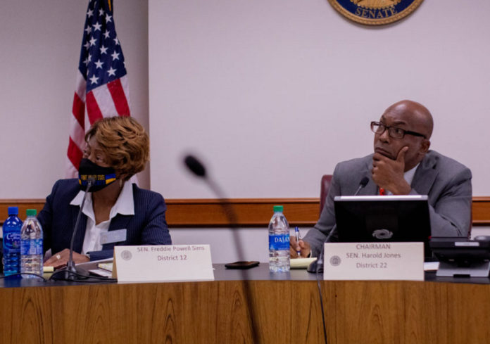 Georgia Food Deserts meeting: Bald black man wearing black frame glasses and blue pin-striped suit with left hand on chin to right of a black woman with blond hair in navy suit and white blouse both sit behind brown wood desk in front of American flag and Senate gold seal
