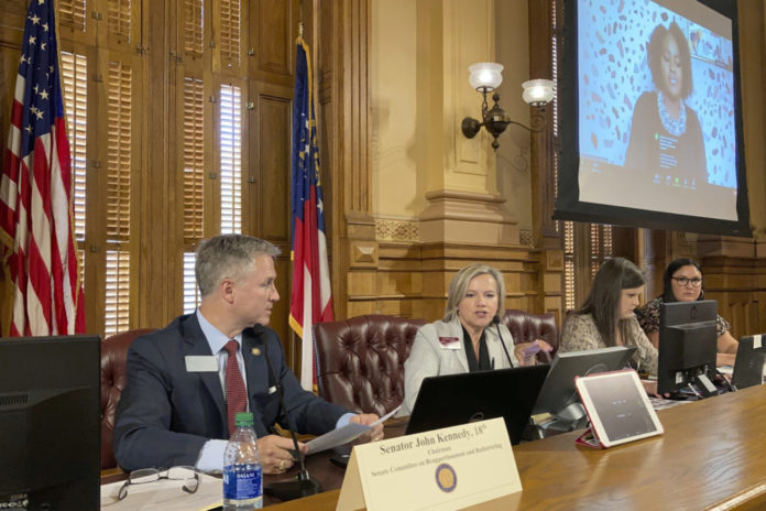 GA Senate redistricting debate: 3 women and 1 man in business attire sit behind a long wood table with laptops and name placards in front of an ornate gold wall, American and Georgia state flags and a large TV monitor with a woman in business suit onscreen.