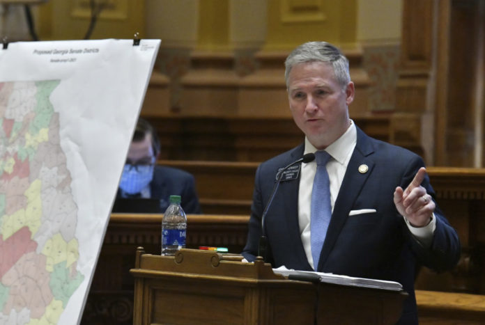 GA Senate redistricting debate: White man with short gray hair in a navy blue suit standing with a blue tie and white shirt pointing his finger to the left standing behind a podium with a georgia color coded redistricting map in front of him