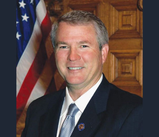 Former state Sen. Ross Tolleson was fondly remembered by his colleagues at the Capitol Senator Ross Tolleson tribute: Smiling gray-haired man iv navy suit and light blue tie stands in front of wood wall next to American flag