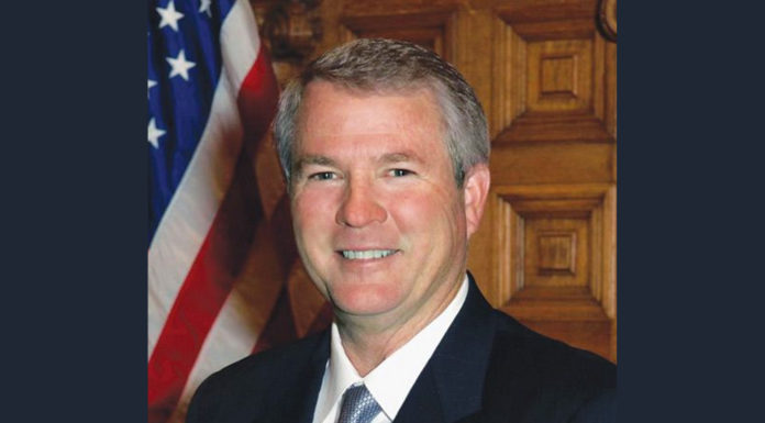 Former state Sen. Ross Tolleson was fondly remembered by his colleagues at the Capitol Senator Ross Tolleson tribute: Smiling gray-haired man iv navy suit and light blue tie stands in front of wood wall next to American flag