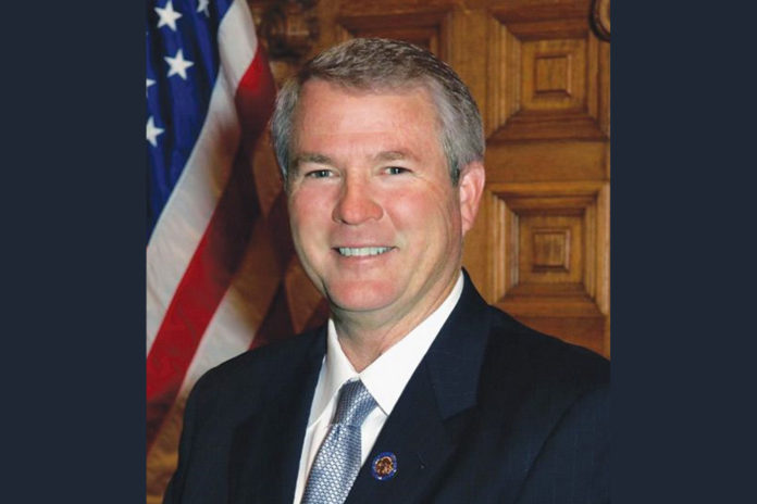 Senator Ross Tolleson tribute: Smiling gray-haired man iv navy suit and light blue tie stands in front of wood wall next to American flag