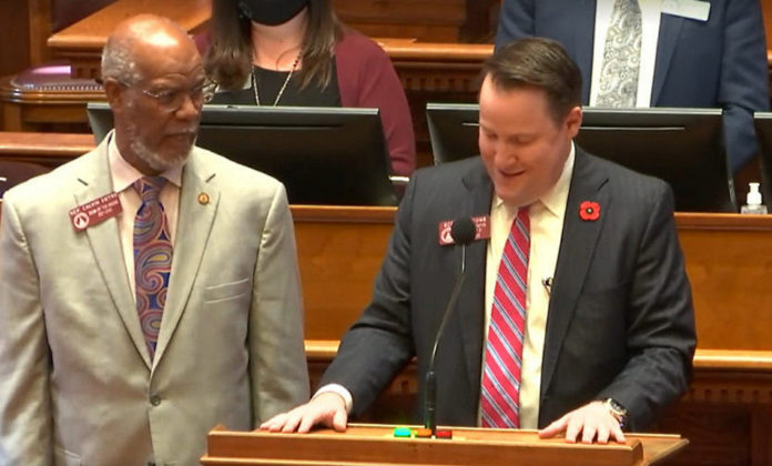Rep. Calvin Smyre remembering Max cleland: A bald black man in Khaki suit next to a short haired white man in a navy suit with a pink tie behind a wooden podium with mic