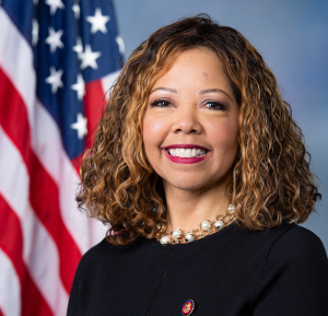 Redistricting map Final: Smiling Black woman with long. curly brown hair, wearing a black top, stands in front of an American flag