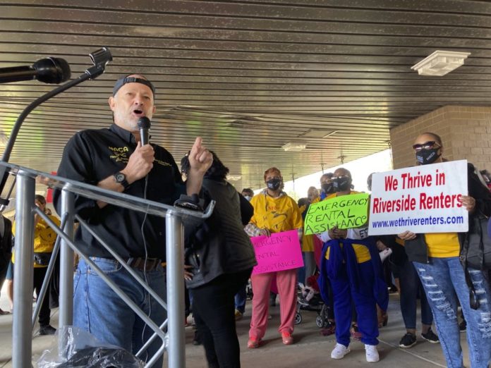 Georgia renters assistance: white man standing at podium holding mic with a black shirt with yellow writing that says NACA in the right corner of shirt surrounded by a group of protesters with homade singsn saying emergency renters assistance and we thrive in riverside renters
