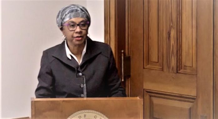 Sickle Cell Committee: Black woman wearing scarf headwrap, glasses and grey suit stands behind brown wood podium speaking into microphone