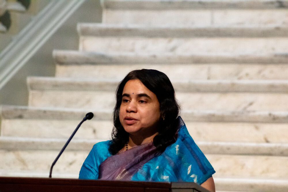 Indian woman wit dark hair and blue and purple dress speaking at a podium.
