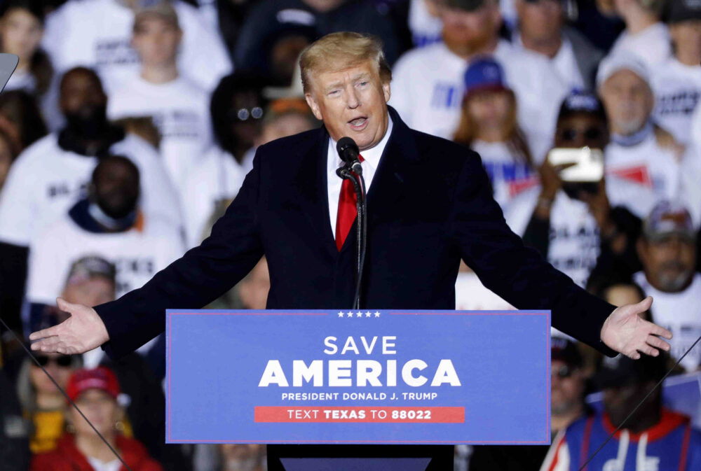 white man with blonde orange hair speaking at a rally at a podium with "Save America. President Donald Trump." " 