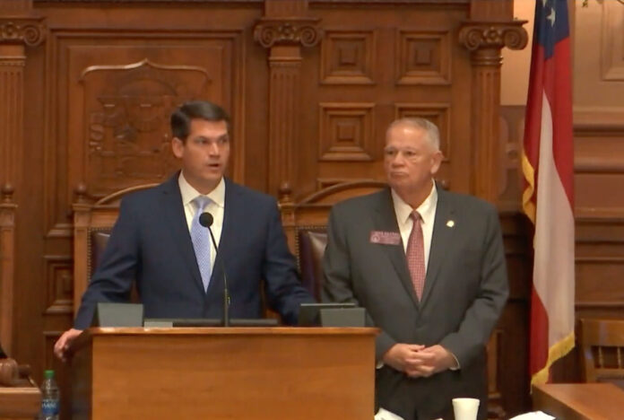 (left) white man standing at podium with navy jacket and blue tie. (right) white man with short white hair with grey jacket, red tie.
