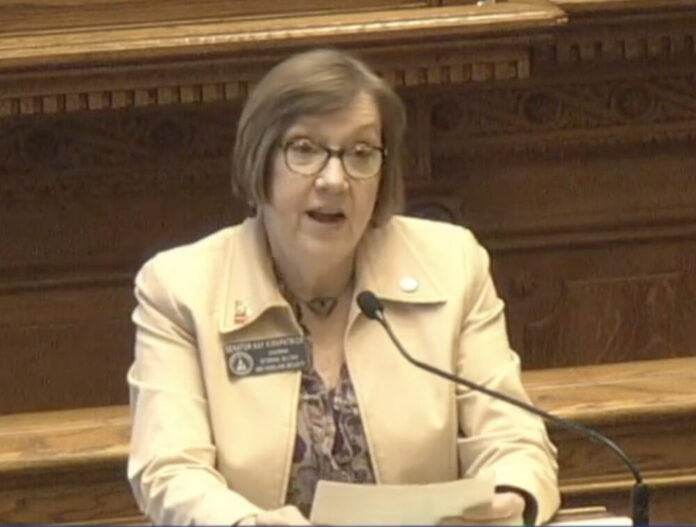 White woman with tan jacket and brown glasses, speaking into microphone