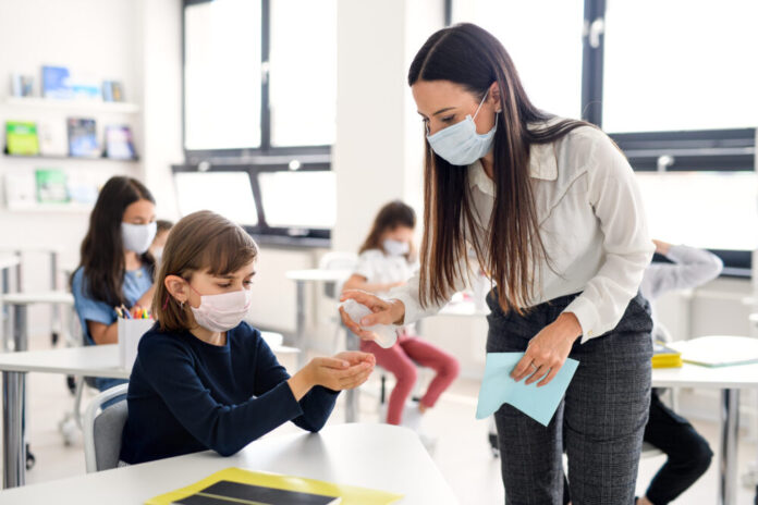 White woman teacher sanitizes her students hand while wearing a mask