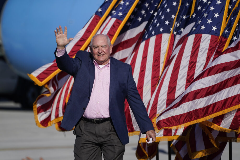 white man walking and waving with American flags blowing in the background