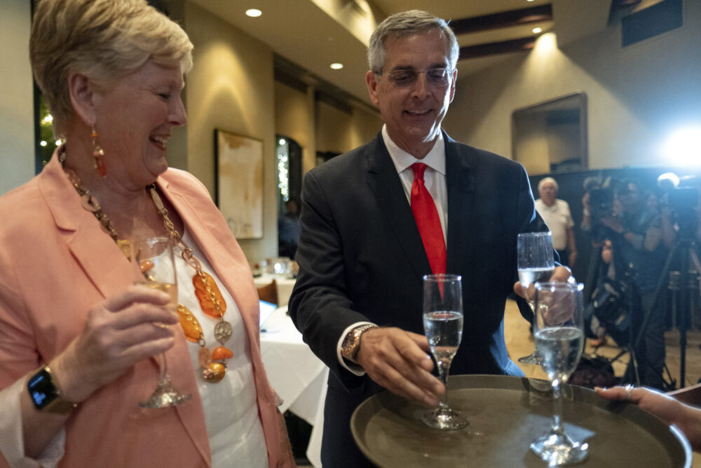 white man in red tie and black jacket holds glass of champagne next to white woman in orange jacket at celebration; Brad Raffensperger