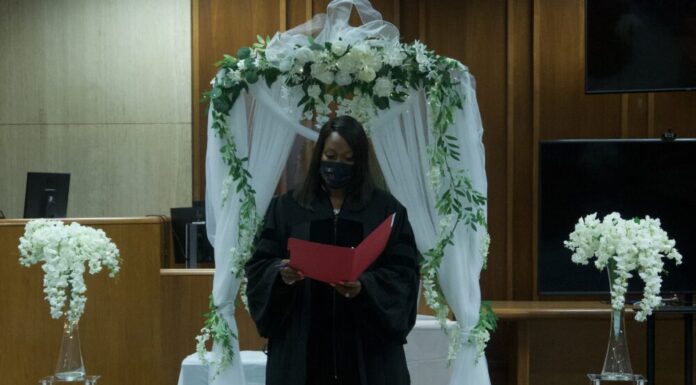 Fulton Probate Court offers free weddings for couples Black woman in judge's robe wearing mask standing in front of white wedding decorations and flowers reds from a red file folder